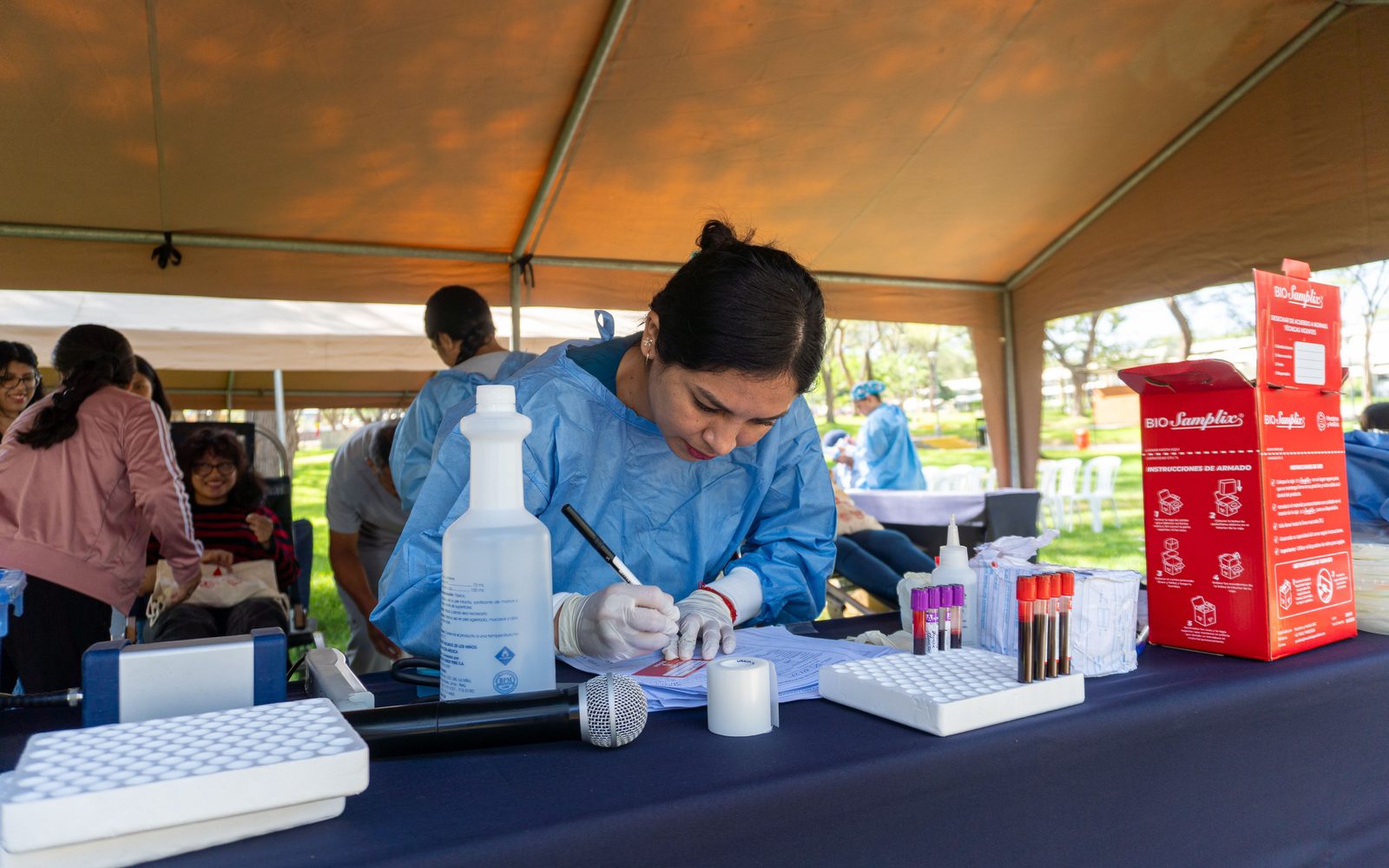 A health worker writing at an outreach table under a canopy at a community clinic day