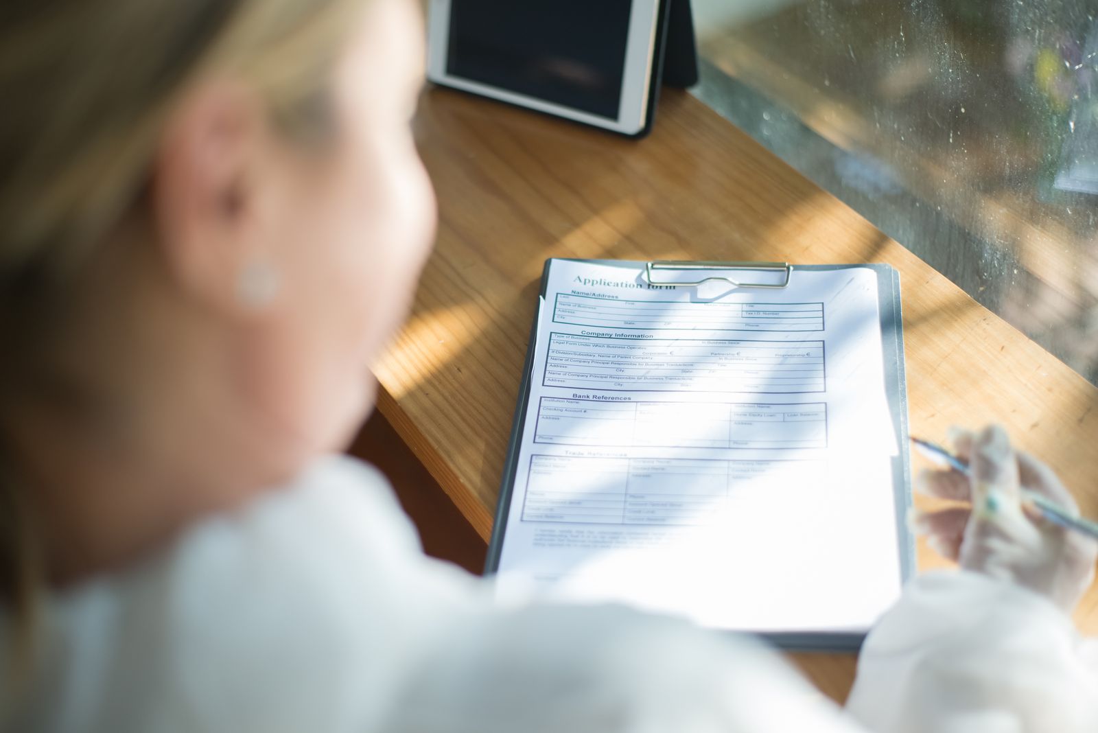 A person filling out an application form with a pen at a sunlit wooden desk