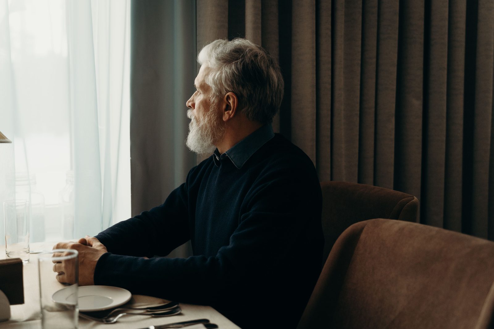 An older man sitting alone at a table with a cup, lit by a window