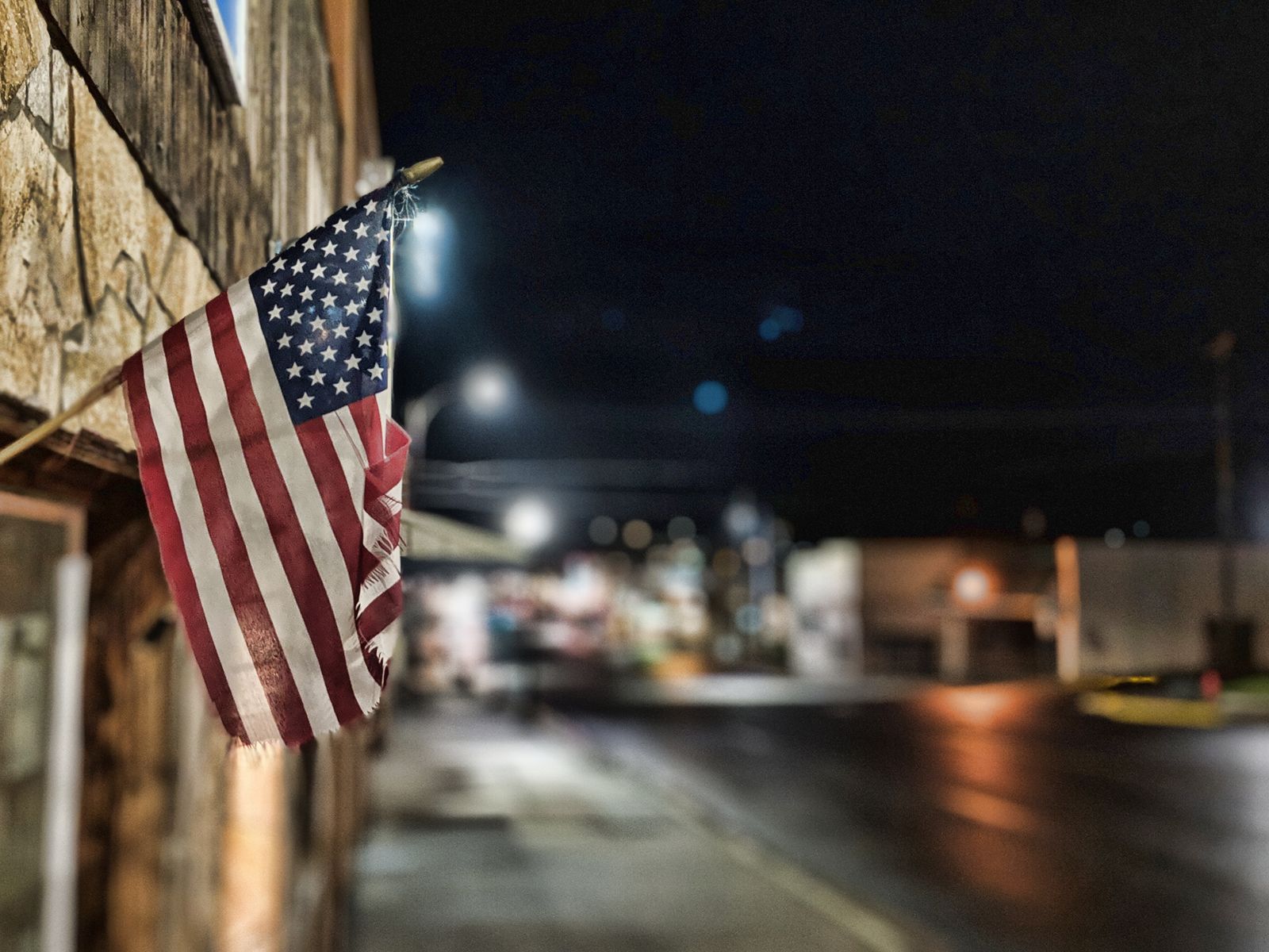 A weathered American flag hanging from a building on a small-town street at night