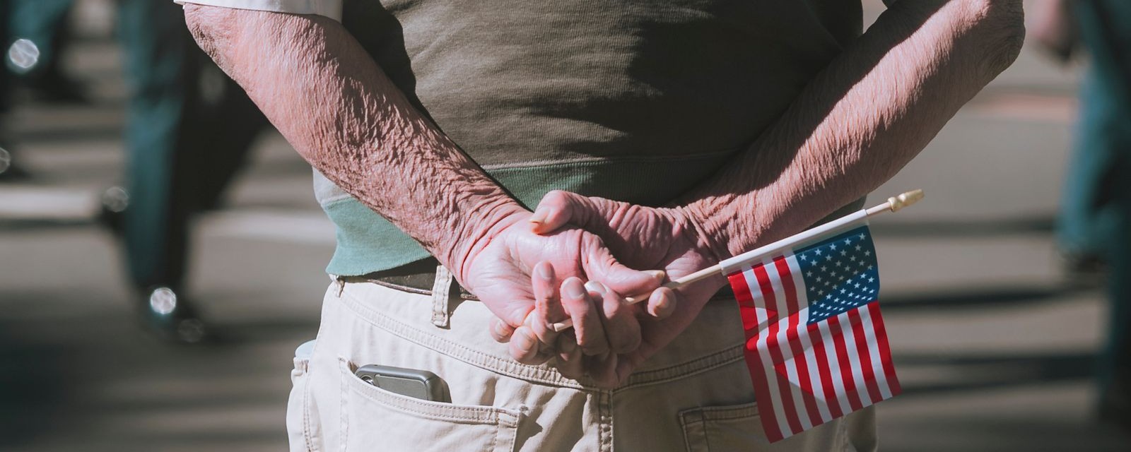 An older veteran's hands clasped behind his back, holding a small American flag at a parade